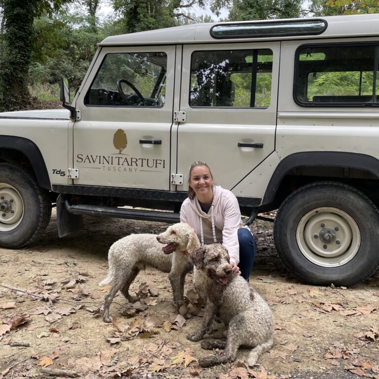 A woman and two truffle hunting dogs are on the ground in front of a jeep labeled Sarvini Tartufi