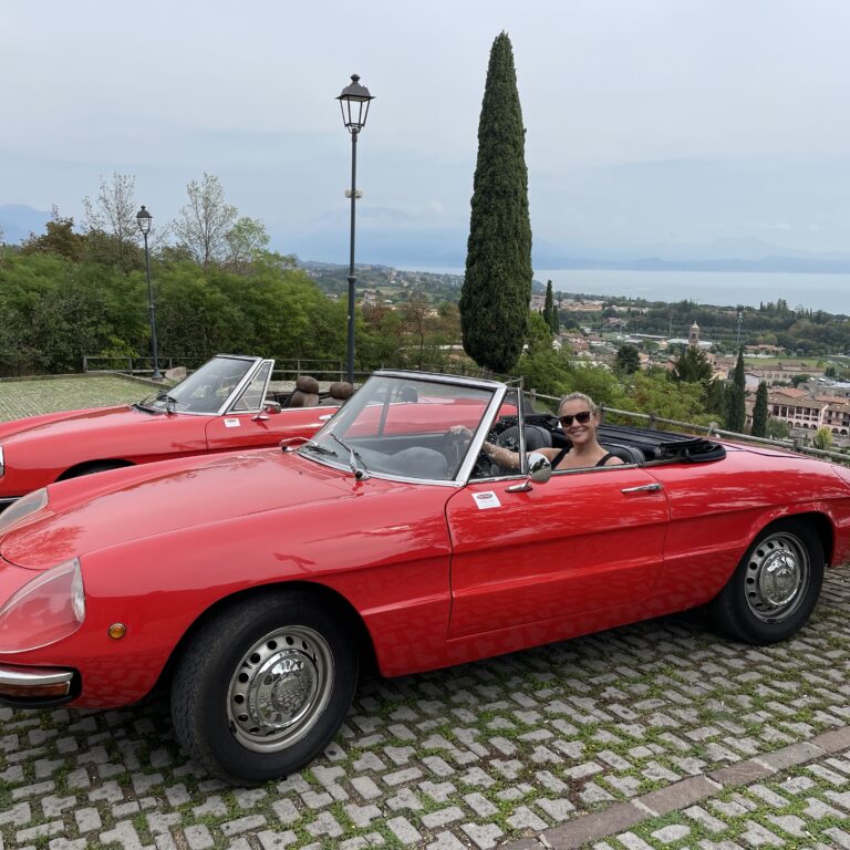 Terry is sitting in a red vintage Alfa Romeo convertible with Lago di Garda in the background.