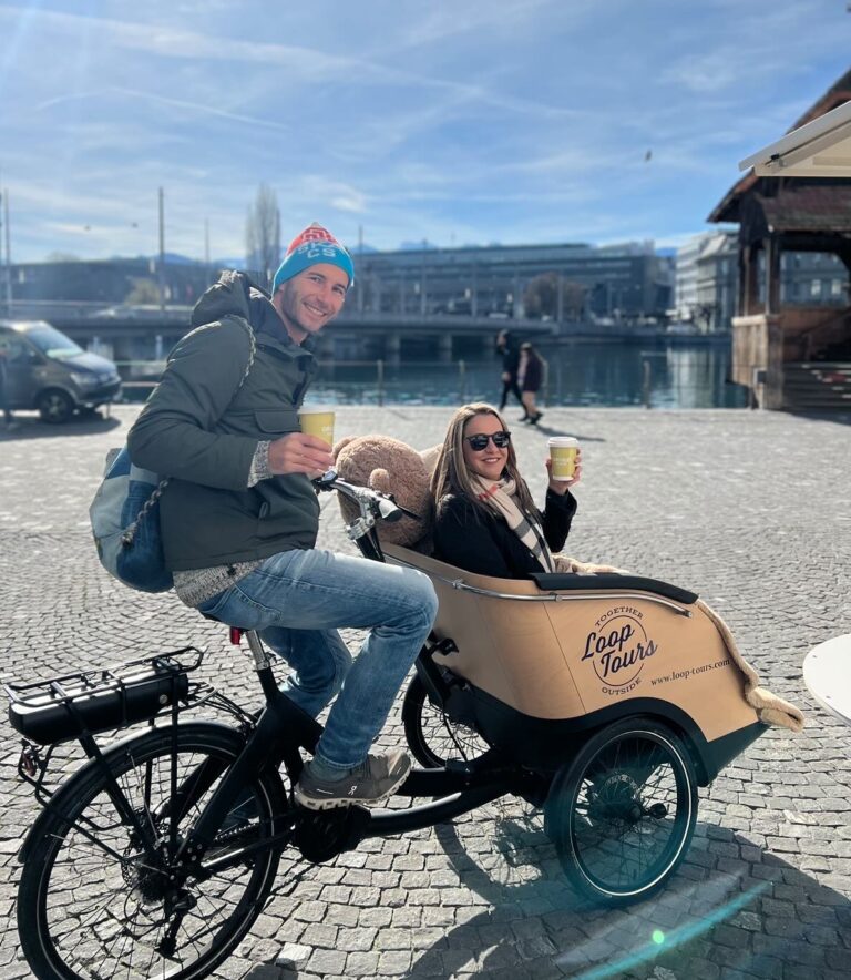 a man is sitting on an electric bike with a cargo piece on the front wheel with a woman sitting in it along with a large bear.
