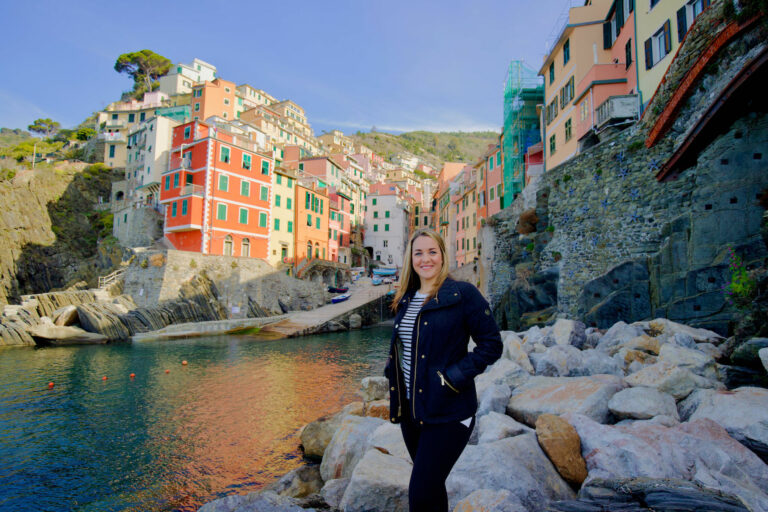 a woman stands on rocks with Riomaggiore in the background during a photo shoot in Cinque Terre Italy