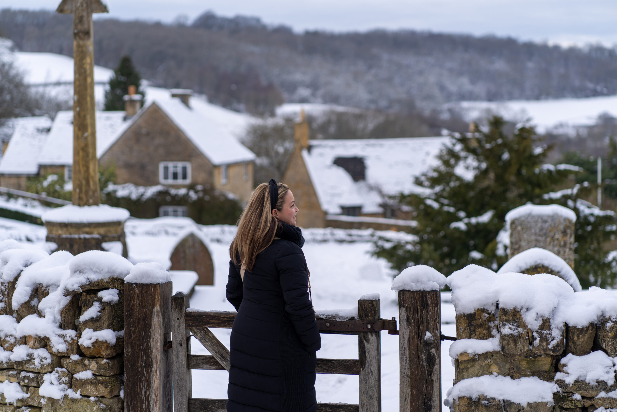 The Cotswolds a woman stands in all black looking out at a snow covered Snowshill in The Cotswolds