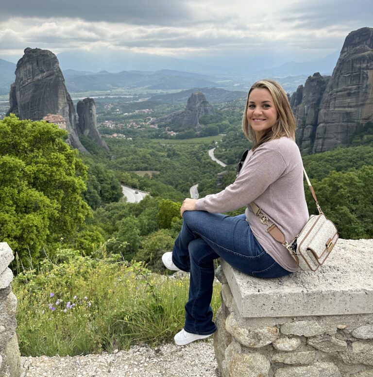 a woman sits on a ledge, legs crossed, with the cliffs of Meteora Greece in the background