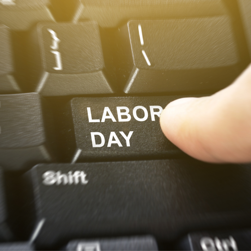 a black computer keyboard has a key that says LABOR DAY and it's being pushed by a finger