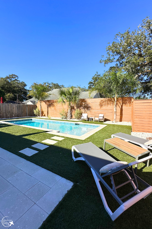 a pool is in a backyard with 3 palm trees, surrounded by pool furniture and astroturf