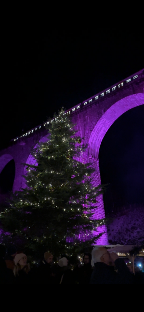 Ravenna Gorge Christmas Market 2 A towering Christmas tree adorned with lights and ornaments in front of the Ravenna Viaduct, illuminated in purple during the Ravenna Gorge Christmas Market in the Black Forest.