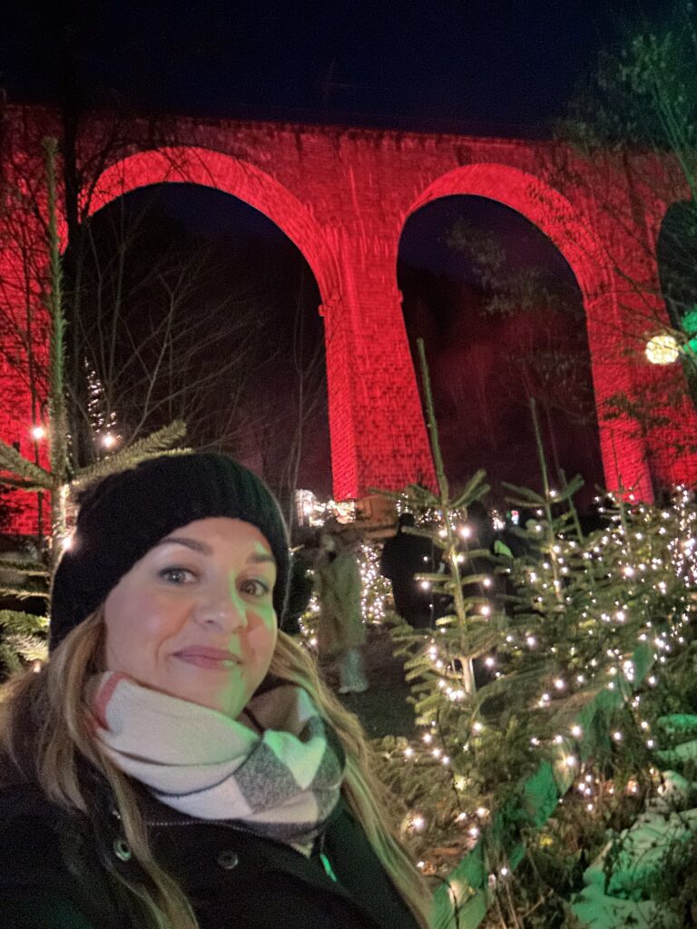 Ravenna Gorge Christmas Market 37 A woman bundled in a warm coat and scarf taking a selfie at the Ravenna Gorge Christmas Market, with the red-lit arches of the Ravenna Viaduct glowing in the background among twinkling lights and trees.