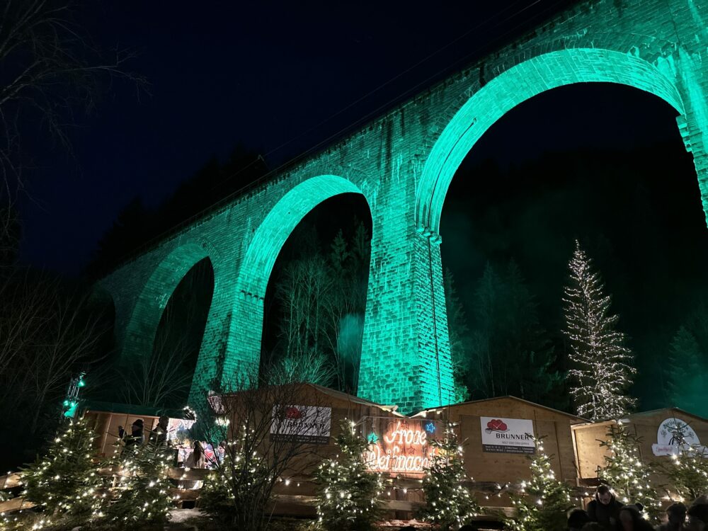 Ravenna Gorge Christmas Market 40 Illuminated Ravenna Viaduct towering over the Ravenna Gorge Christmas Market in the Black Forest, Germany, with festive wooden stalls and twinkling lights surrounded by Christmas trees.