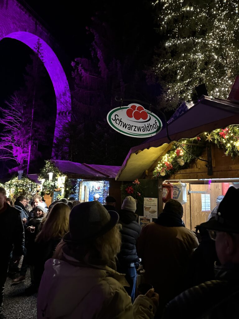 Ravenna Gorge Christmas Market 50 Visitors queuing at a festive food stall at the Ravenna Gorge Christmas Market, with the Schwarzaldhof sign and purple-lit Ravenna Viaduct in the background.