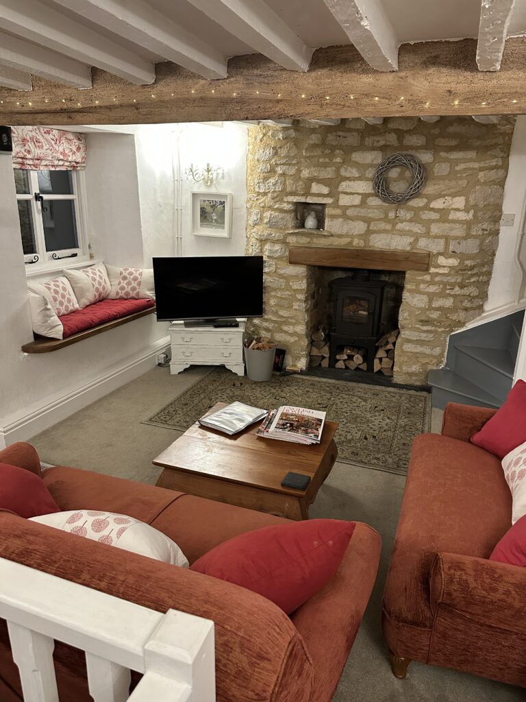 Chalford Cottage Cotswolds 17 Charming living room at Minnow Cottage with a picturesque window seat, a stone fireplace, and rustic beams, offering a perfect blend of comfort and charm.