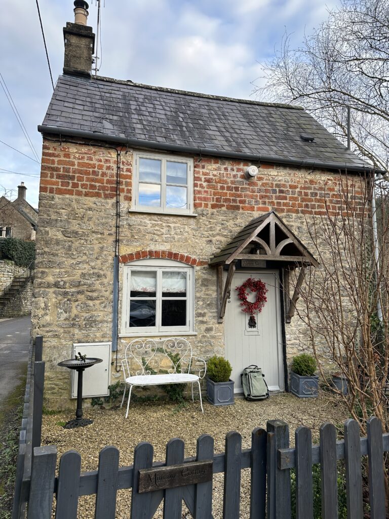 Chalford Cottage Cotswolds 23 Charming exterior of Minnow Cottage in Chalford, Cotswolds, with a red berry wreath on the door, white bench, and rustic stone facade, exuding cozy countryside charm.