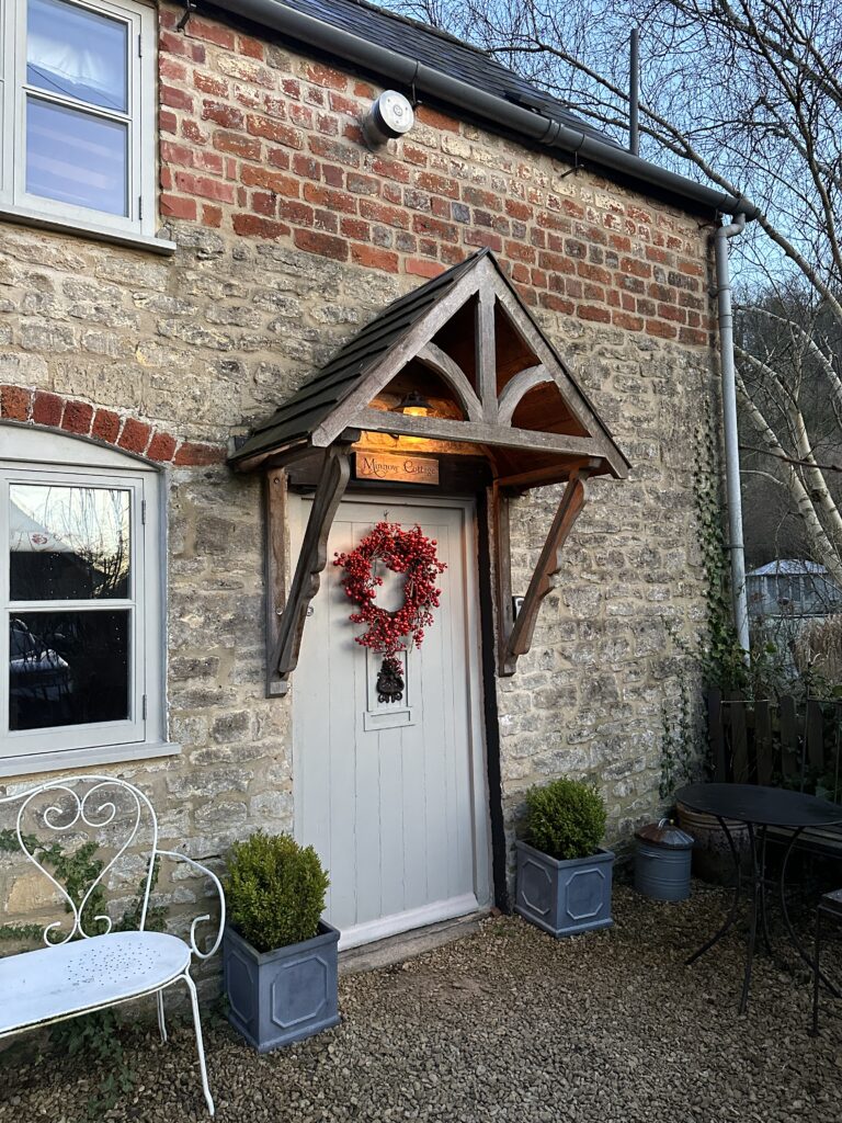 Chalford Cottage Cotswolds 3 Cozy entrance of Minnow Cottage in Chalford, Cotswolds, featuring a charming wooden awning, a festive wreath on the door, and a white bench for a welcoming vibe.