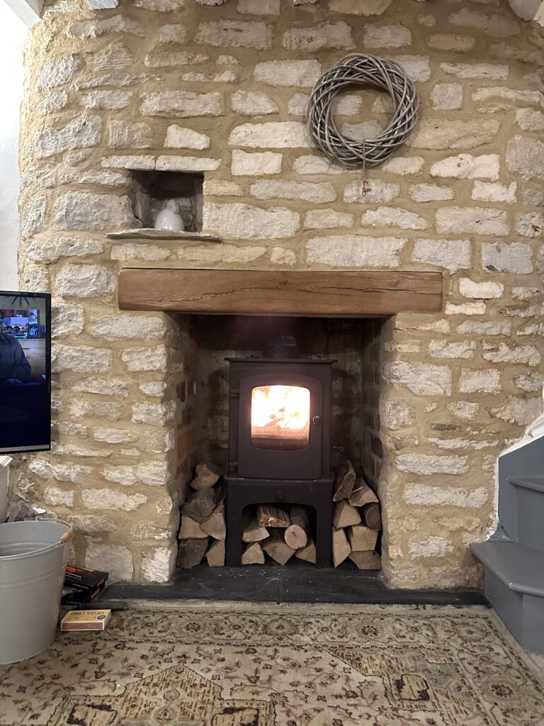 Chalford Cottage Cotswolds 30 Cozy wood-burning stove at Minnow Cottage in the Cotswolds, nestled in a rustic stone fireplace with neatly stacked firewood, creating a warm and inviting ambiance.