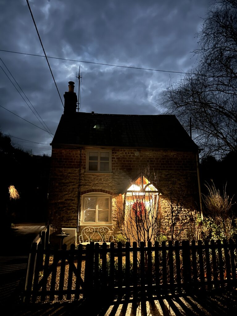 Chalford Cottage Cotswolds 31 Enchanting nighttime view of Minnow Cottage in Chalford, Cotswolds, with warm glowing lights illuminating the entrance, set against a dramatic cloudy sky.