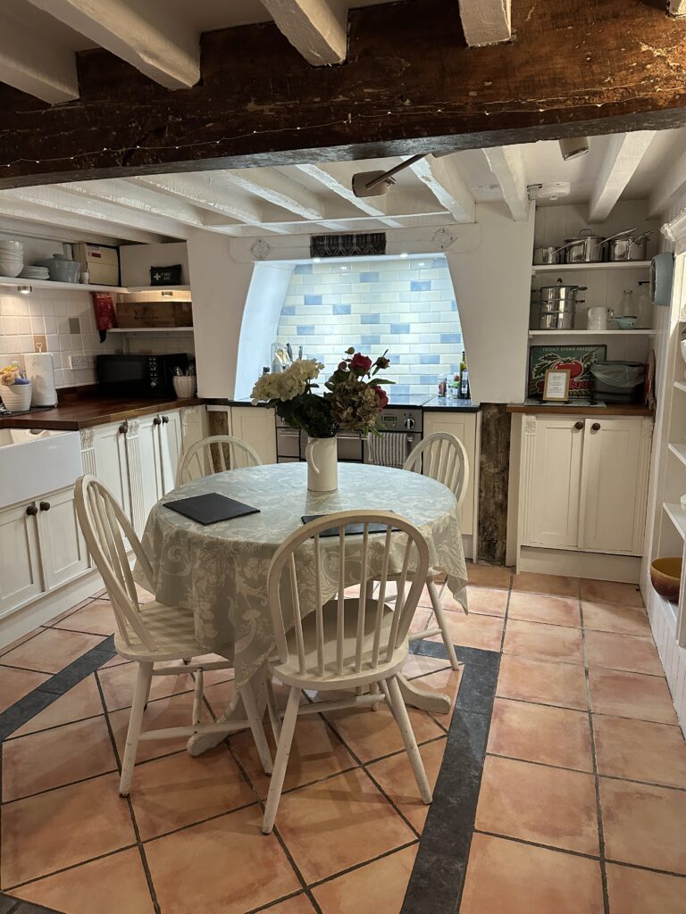 Chalford Cottage Cotswolds 6 Rustic kitchen at Minnow Cottage in the Cotswolds with exposed beams, tiled floors, a round dining table, and modern amenities for a cozy countryside stay.