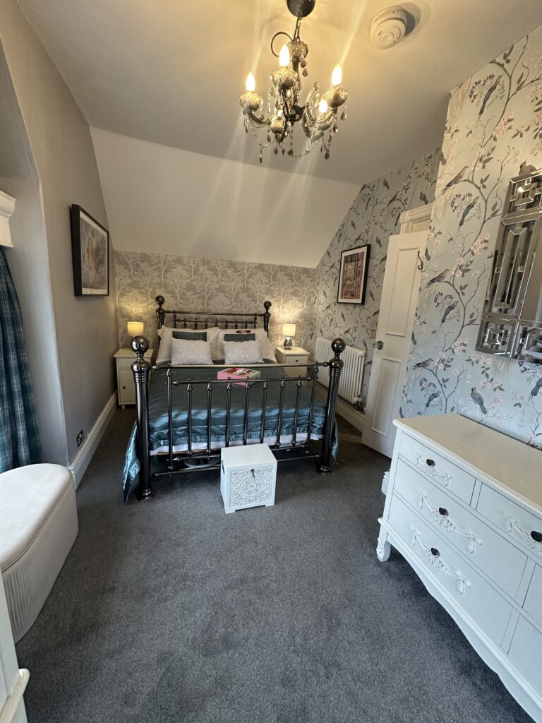 Stratford Upon Avon Penthouse 1 Elegant bedroom in a Stratford-Upon-Avon penthouse, featuring a vintage-style metal bed, floral wallpaper, and a sparkling chandelier for a cozy, luxurious stay.