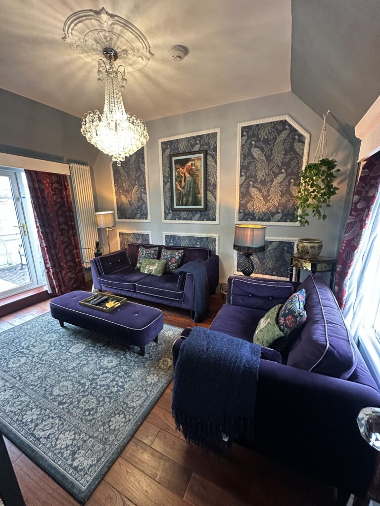 Stratford Upon Avon Penthouse 11 Luxurious living room in a Stratford-Upon-Avon penthouse, featuring rich purple seating, elegant wall decor, and a sparkling crystal chandelier.