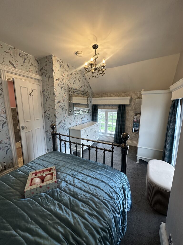 Stratford Upon Avon Penthouse 2 View of the charming bedroom in a Stratford-Upon-Avon penthouse, highlighting a plush teal comforter, decorative dresser, and natural light from large windows.