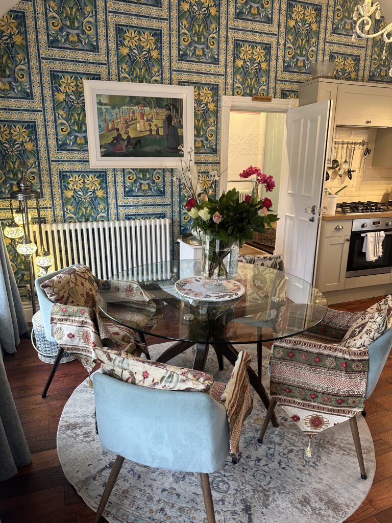 Stratford Upon Avon Penthouse 6 Close-up of a charming dining setup in a Stratford-Upon-Avon penthouse, featuring a glass-top table, fresh flowers, and intricate wallpaper as a backdrop.