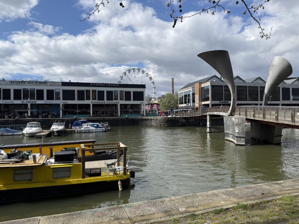 IMG_8008 Bristol Harborside with boats on the water, modern buildings, and the distinctive Pero’s Bridge with horn-shaped sculptures under a partly cloudy sky