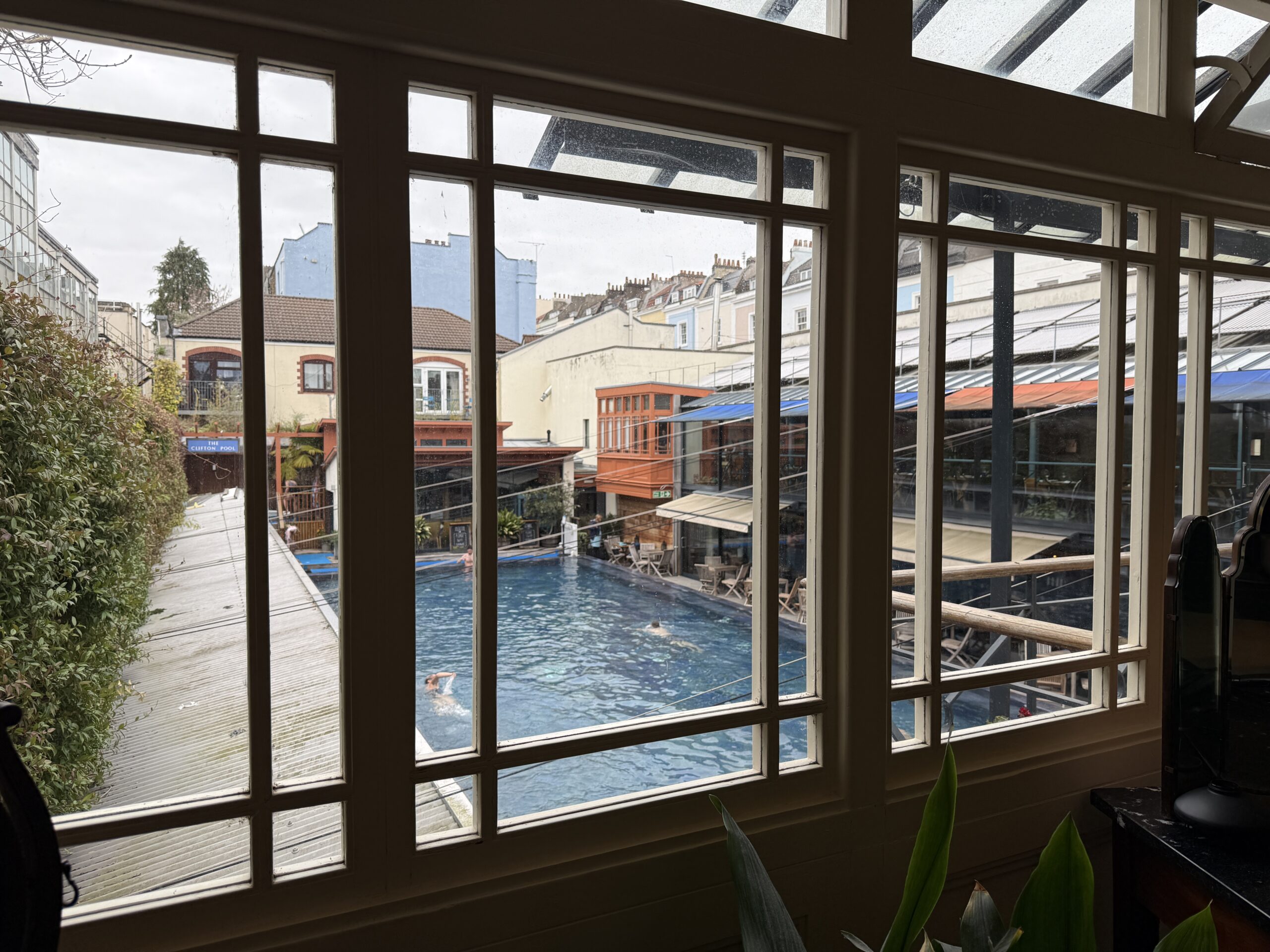 Bristol Lido outdoor pool seen through the interior windows of the Victorian bathhouse in Clifton with swimmers in the heated pool and Bristol rooftops in the background