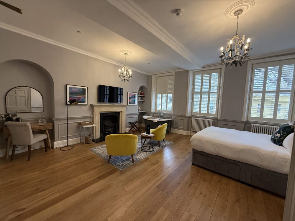 Spacious boutique hotel room at Eight Hotel Bath with oak floors, twin chandeliers, yellow velvet armchairs, original fireplace, and a freestanding bathtub by the window