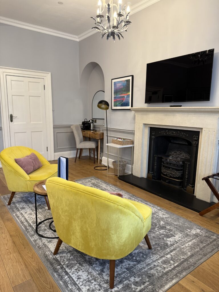 Boutique hotel sitting area at Eight Hotel Bath with two yellow velvet armchairs, original Victorian cast iron fireplace, chandelier, and Georgian cornicing