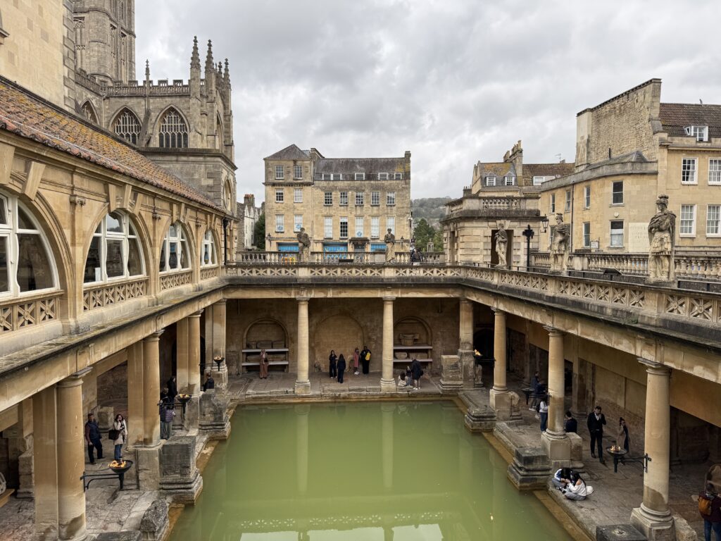 Roman Baths in Bath, England with green thermal water pool surrounded by stone columns and historic buildings under a cloudy sky