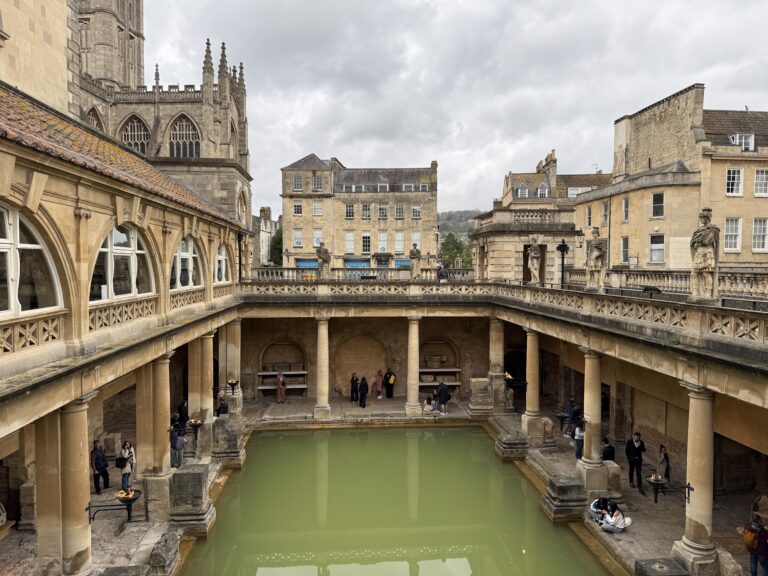 Roman Baths in Bath, England with green thermal water pool surrounded by stone columns and historic buildings under a cloudy sky