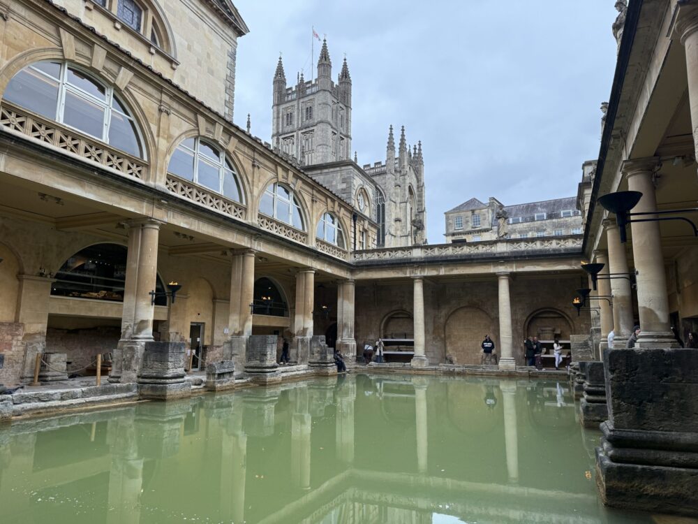 View across the Great Bath at the Roman Baths in Bath England showing the mineral-green thermal pool, surrounding colonnades, and the Gothic tower of Bath Abbey rising above the ancient ruins