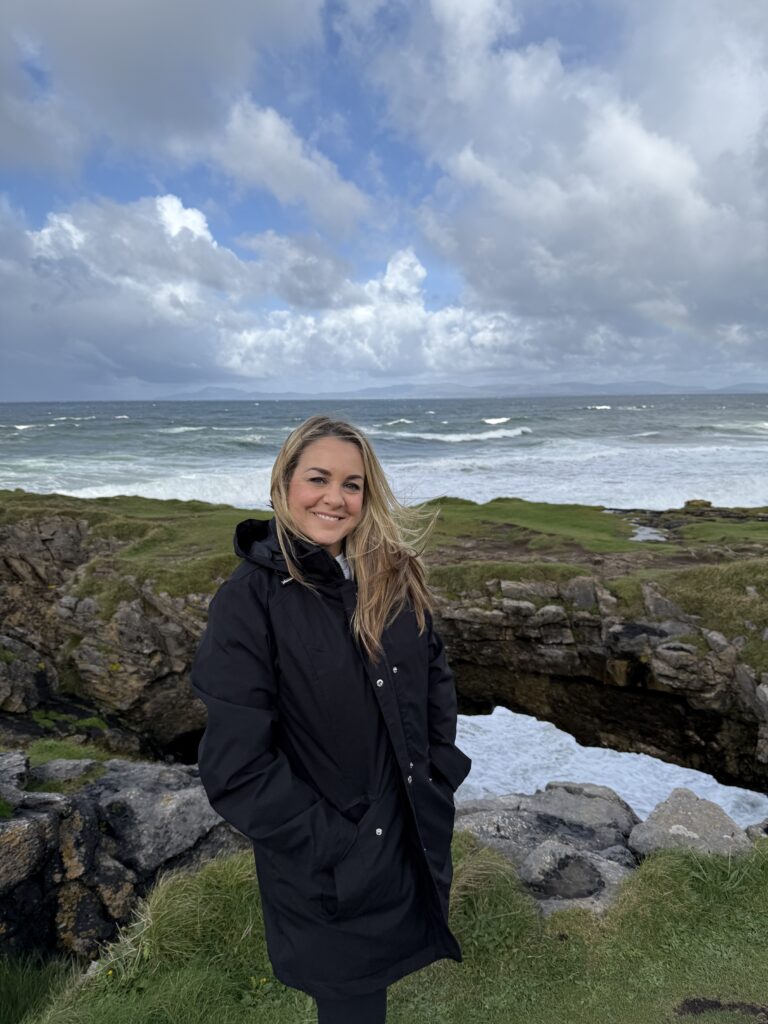 A smiling woman with long blonde hair stands in a black jacket before a rocky coastline and a turbulent ocean.