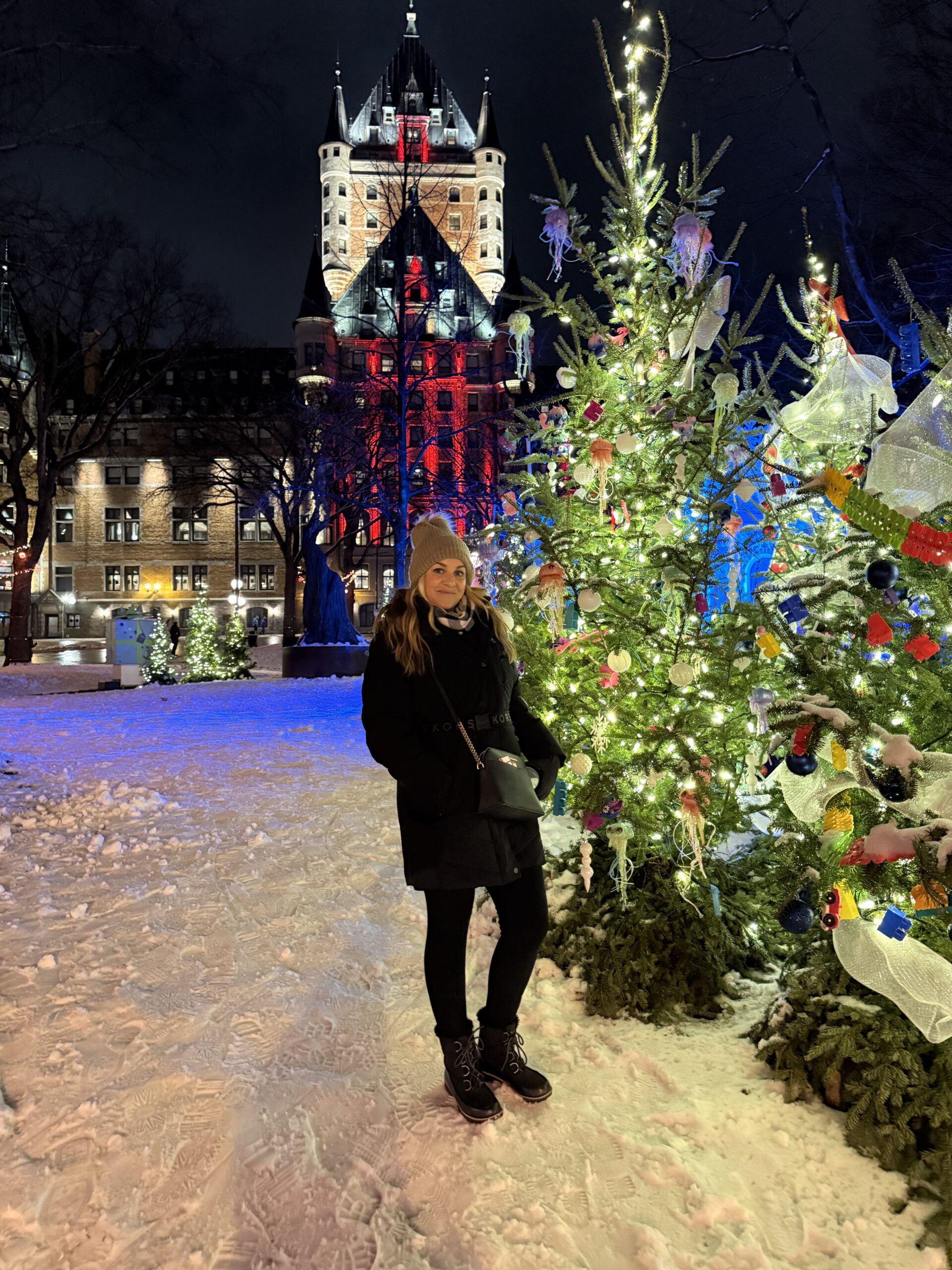 A person in a winter coat and hat stands in a snowy park at night next to a decorated Christmas tree near a large building.