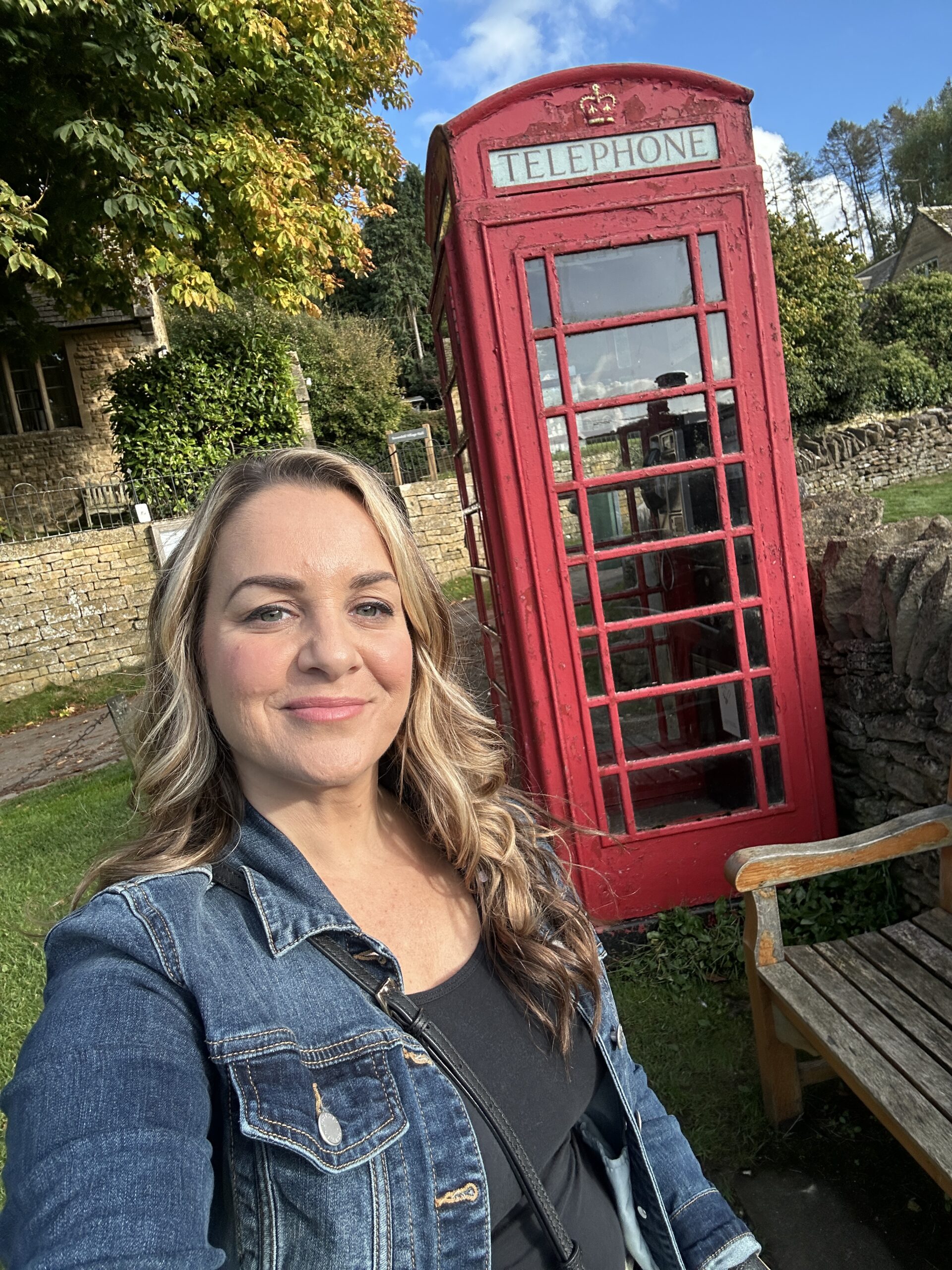 A woman with long blonde hair wearing a denim jacket takes a selfie in front of a red telephone box.