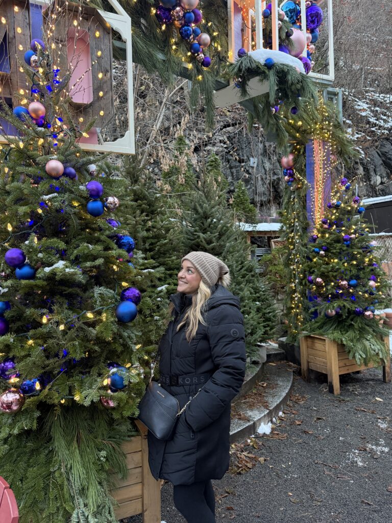 A woman in a winter coat and beanie stands smiling beside a Christmas tree decorated with blue and pink ornaments.